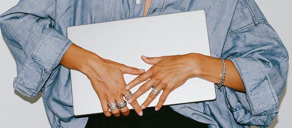 Hands wearing rings resting on a closed silver laptop, in an oversized denim shirt.