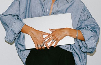 Hands wearing rings resting on a closed silver laptop, in an oversized denim shirt.