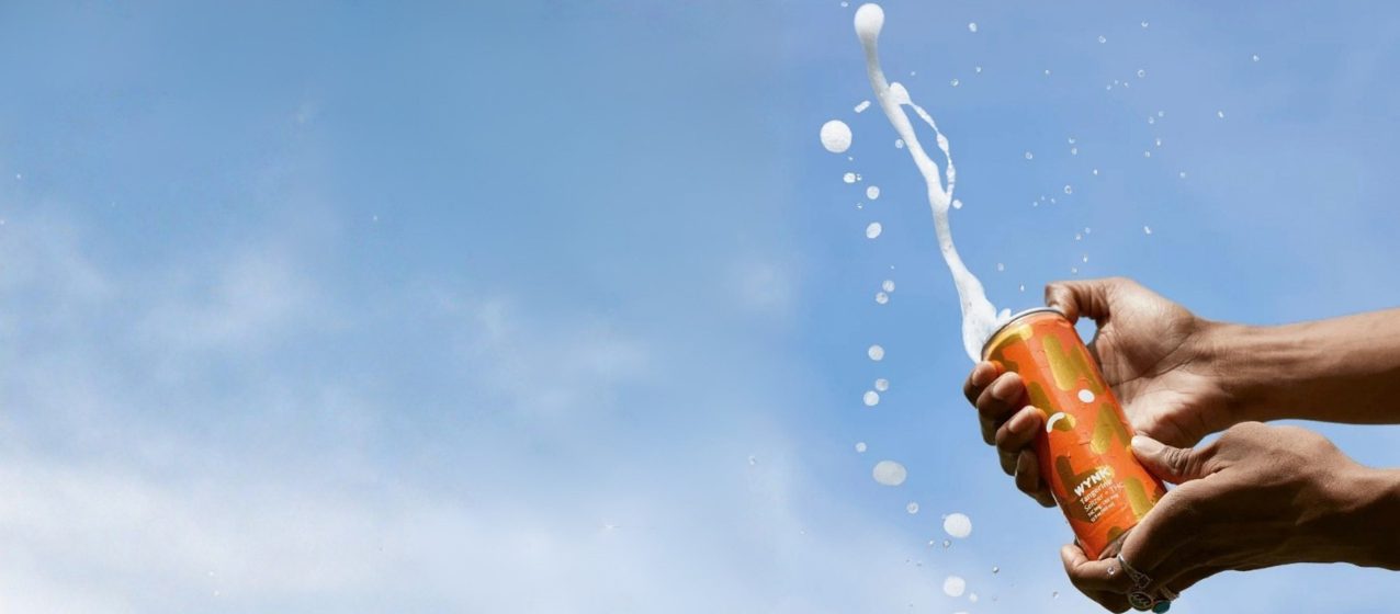 A hand holding an orange can against a blue sky as the drink sprays out of the top.