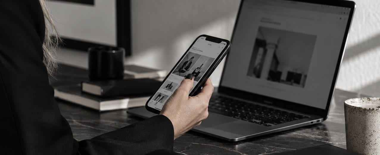 A hand holding a phone showing a social media profile in front of an open laptop on a dark desk.