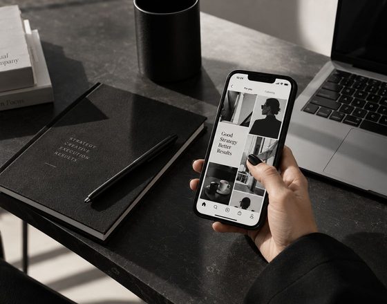 A hand holding a phone showing a grid of ad creatives above a dark desk with a notebook and a laptop.