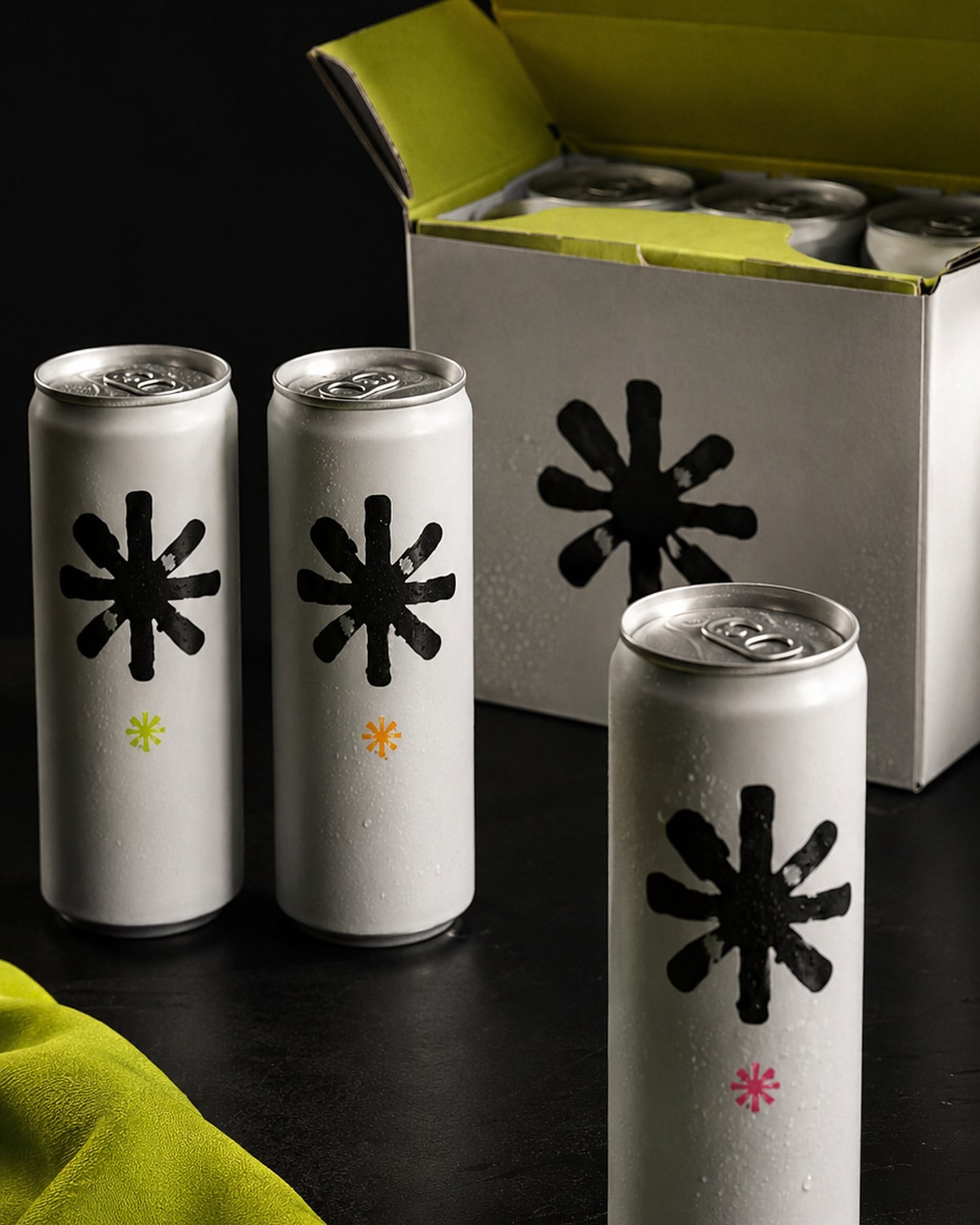 Three white beverage cans with asterisk marks and colored flavor dots beside an open lime-lined multipack carton