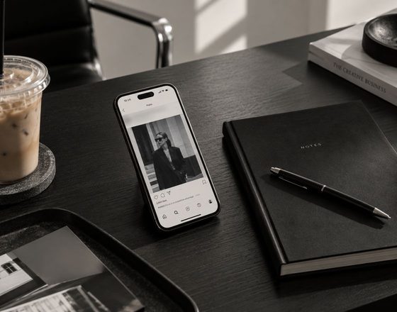 A phone showing a social media profile on a dark desk next to a notebook, a pen, and an iced coffee.